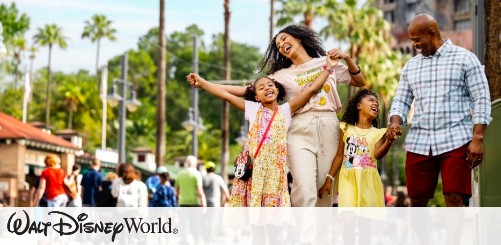 A joyful family of four walks hand in hand at a theme park, surrounded by palm trees and other visitors. The children wear colorful outfits with cartoon characters, while the parents are dressed casually. The sun is shining, creating a vibrant and lively atmosphere.