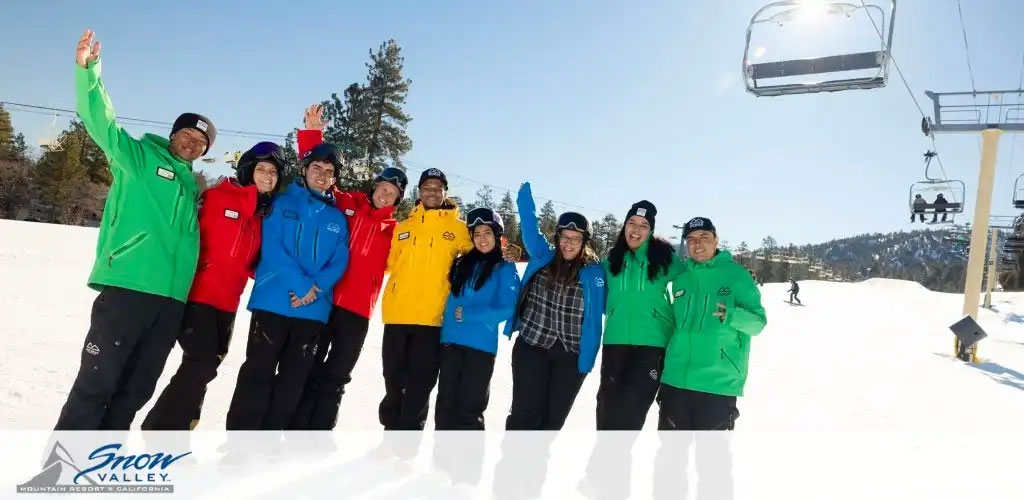 This image depicts a cheerful group of nine ski resort staff members standing on a snowy slope under a bright blue sky. On the left, a ski lift chair hovers off the ground, with several empty chairs visible in the line, indicating a peaceful day at the resort. The staff, a mix of men and women, are dressed in colorful uniforms—vibrant red, blue, yellow, and various shades of green jackets with contrasting snow pants. They are smiling widely and playfully waving or raising their hands to the camera, creating a welcoming and friendly atmosphere. Each jacket is adorned with a name tag, and they wear either winter hats or helmets, suggesting readiness for safe and fun activities on the slopes. The snow beneath their feet is groomed, with some ski tracks visible, and a few individuals can be seen in the background enjoying the ski area. The Snow Valley Mountain Resort logo is prominently displayed in the lower left corner, indicating the location in California.

Discover the joy of the slopes and lock in your mountain adventure with the lowest prices on tickets—it's all downhill from here when it comes to savings at GreatWorkPerks.com!