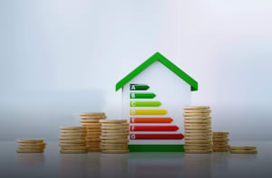 Hausmodell mit Energieeffizienzskala von A bis G zwischen Münzstapeln als Symbol für Energiekosten und Sanierung