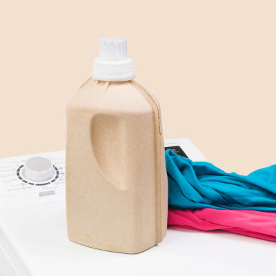 Wäsche im Hintergrund. Toplader-Waschmaschine mit natürlichem Bio-Waschmittel und bunter Kleidung, isoliert vor einem beigen Hintergrund.