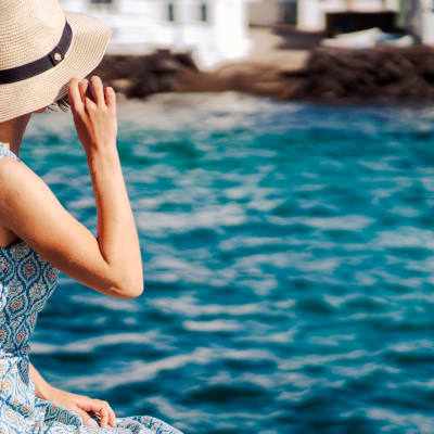 Ein Mädchen in einem wunderschönen Kleid hält einen eleganten Hut in den Händen und genießt ihre Freizeit, während sie sich am Meer entspannt. Eine Dame steht mit dem Rücken zum Ufer und blickt auf das Meer und die Stadt am Horizont.