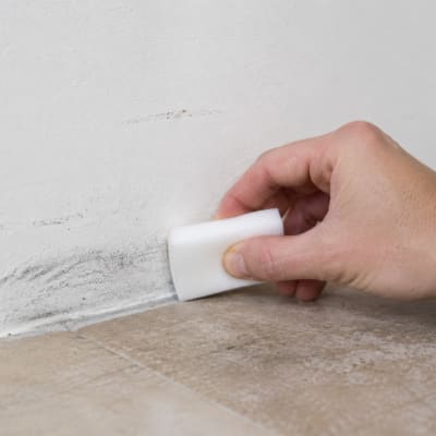 Eine Hand reinigt mit einem weißen Schmutzradierer dunkle Abriebspuren an einer weißen Wand direkt über einem Fliesenboden. Die Nahaufnahme zeigt den Reinigungsvorgang