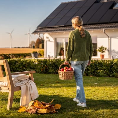 Frau im grünen Strickpullover trägt einen Korb mit Äpfeln durch einen herbstlichen Garten, dahinter ein Einfamilienhaus mit Solaranlage auf dem Dach und Windräder am Horizont