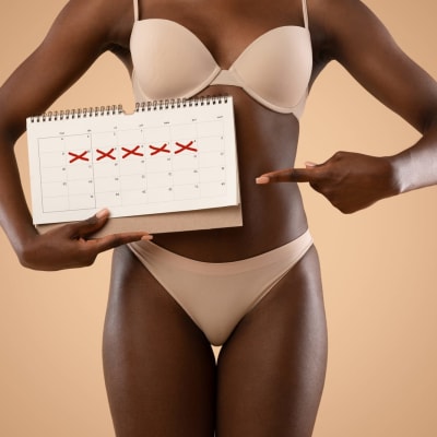 Eine junge afroamerikanische Frau in beiger Unterwäsche zeigt auf einen Kalender mit markierten Tagen.