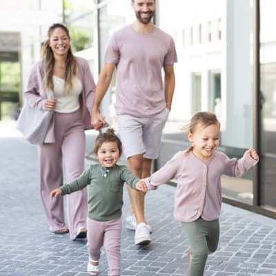 Fröhliche Familie spaziert durch eine Stadtgasse – Eltern und zwei Kinder in aufeinander abgestimmter Kleidung in Rosa- und Grüntönen von ENGEL Natur.