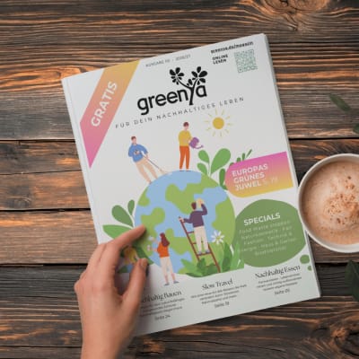 Greenya Magazin wird in der Hand gehalten von jemandem mit Kaffee daneben auf Holztisch. 