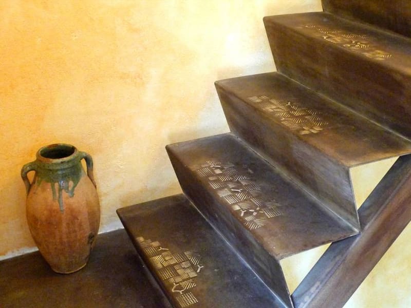 Stahltreppe mit eingeschmiedeten Strukturen in den Stufen vor einer gelben Wand, daneben ein Tonkrug | © Hofschmiede Dahlem (Fotos: Mike Wolff, Torsten Theel)