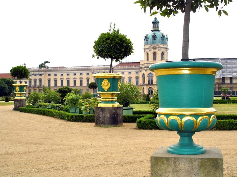 Grün und golden gefasste gusseiserne Vasen auf Sockeln im Schlosspark Charlottenburg, im Hintergrund das Schloss | © Hofschmiede Dahlem (Fotos: Mike Wolff, Torsten Theel)