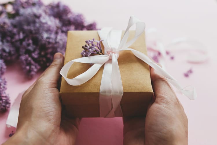 Hände halten ein kleines Päckchen mit weißer Schleife und Trockenblume als Dekoration