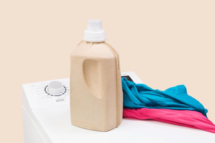 Wäsche im Hintergrund. Toplader-Waschmaschine mit natürlichem Bio-Waschmittel und bunter Kleidung, isoliert vor einem beigen Hintergrund.