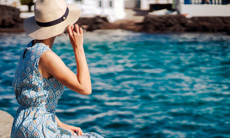 Ein Mädchen in einem wunderschönen Kleid hält einen eleganten Hut in den Händen und genießt ihre Freizeit, während sie sich am Meer entspannt. Eine Dame steht mit dem Rücken zum Ufer und blickt auf das Meer und die Stadt am Horizont. null