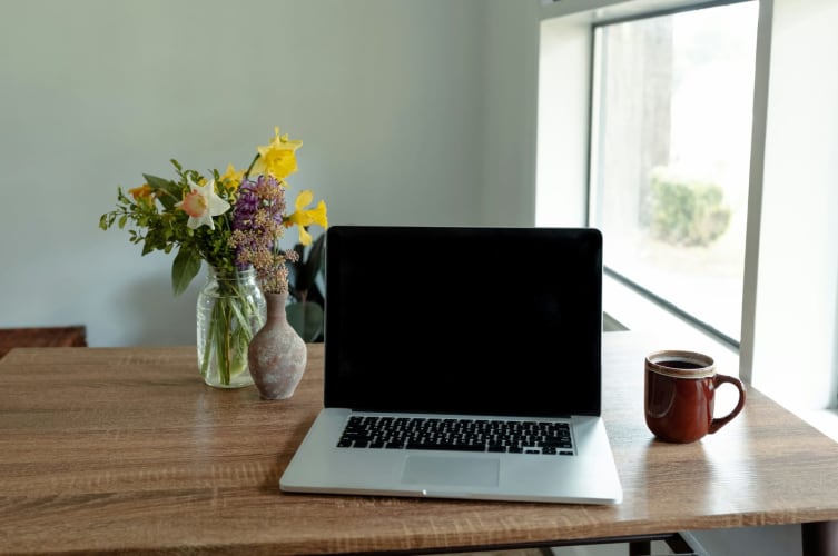 Aufgeklappter Laptop auf einem Holztisch neben Blumenstrauß und Tasse am Fenster © Sandra Seitamaa / Unsplash+
