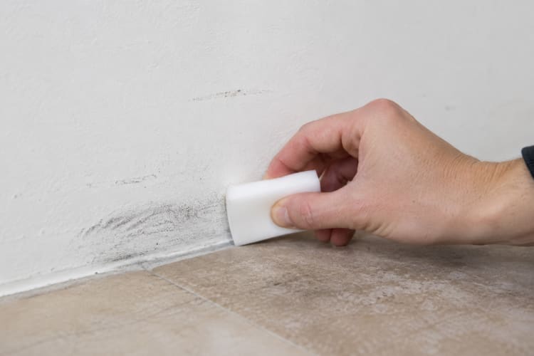 Eine Hand reinigt mit einem weißen Schmutzradierer dunkle Abriebspuren an einer weißen Wand direkt über einem Fliesenboden. Die Nahaufnahme zeigt den Reinigungsvorgang