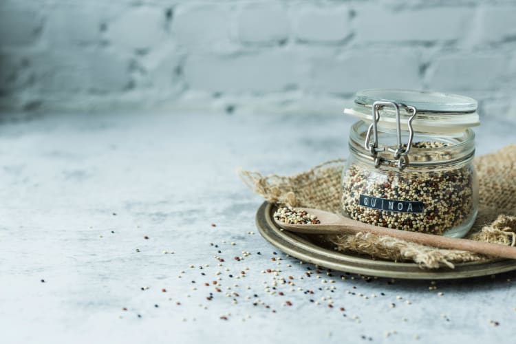 Ein kleines Weckglas mit buntem Quinoa drin und der Aufschrift Quinoa und einem kleinen Holzlöffel auf dem Quinoa liegt  null