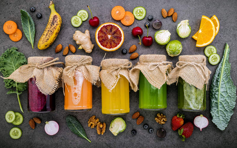 Farbenfrohe, gesunde Smoothies und Säfte in Flaschen mit frischen tropischen Früchten und Superfoods vor einem dunklen Stein-Hintergrund mit Platz für Text.