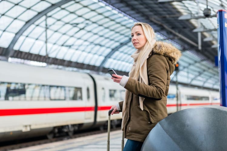Frau am Bahnhof nutzt Fahrplan-App auf ihrem Handy, während ein moderner Zug einfährt. null