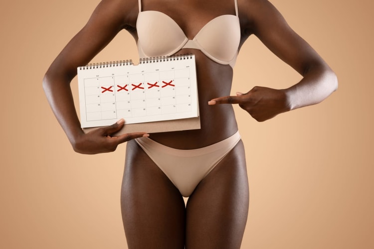 Eine junge afroamerikanische Frau in beiger Unterwäsche zeigt auf einen Kalender mit markierten Tagen. null