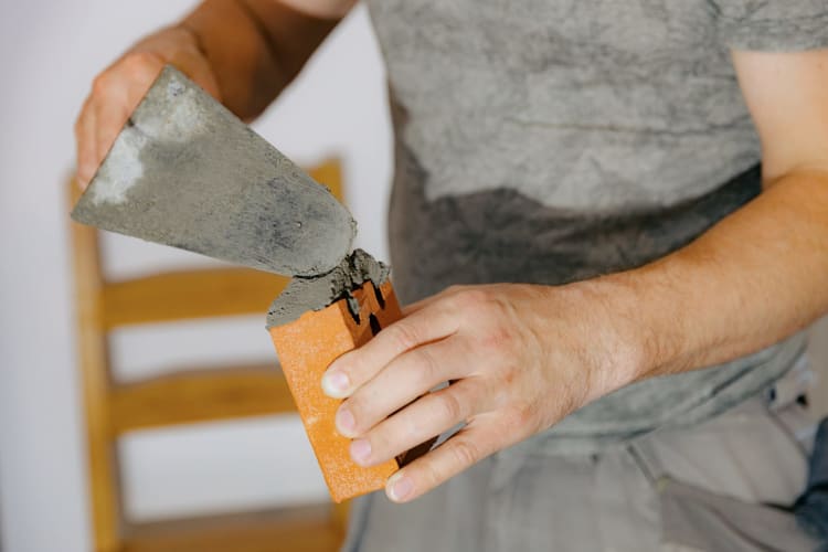 Ein Handwerker, der mit einer Kelle frischen Zementmörtel auf einen Ziegelstein aufträgt, um den Schornstein eines Kamins in einem Raum zu errichten; Nahaufnahme aus der Seitenansicht mit selektiver Schärfe. Konzept für sauberes Mauerwerk, nachhaltige Hausrenovierung.