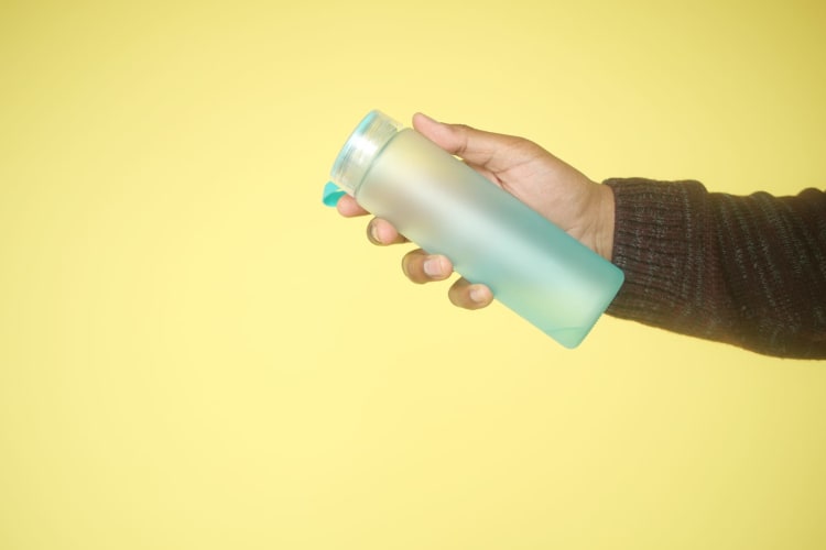 Nahaufnahme einer Hand, die eine blaue wiederverwendbare Wasserflasche vor einem leuchtend gelben Hintergrund hält.