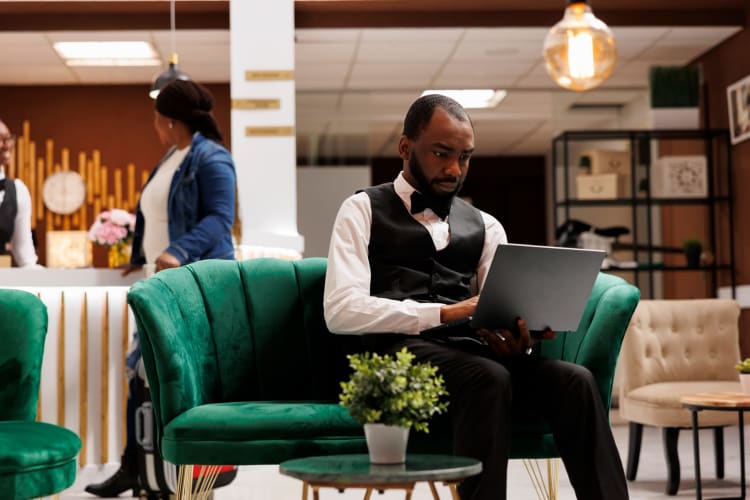 Der Hotelmanager sitzt in der Lobby, arbeitet an seinem Laptop, schaut auf den Bildschirm und behält den Überblick über die Buchungen.