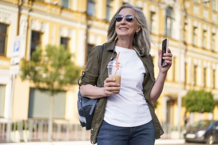 reife europäische Frau mit grauem Haar und Sonnenbrille spaziert mit einem Getränk und einem Smartphone durch die Stadt. Gesunde Langlebigkeit null