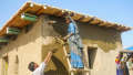 Woman up ladder putting on render, man handing up more render