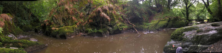 Panorama of steep sided small river from the waterfall, trees to the left, bridge to the right