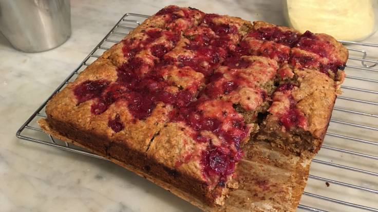 Square cake sliced on cooling rack, red fruit on top, piece missing