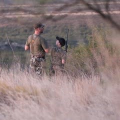father and son carrying a shotgun through tall grass at dusk