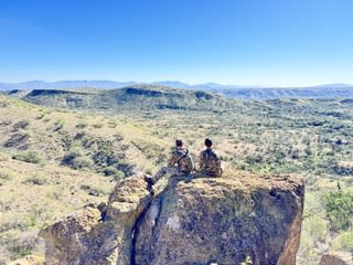 two hunters in camo sitting on a rocky point overlooking a wide desert valley