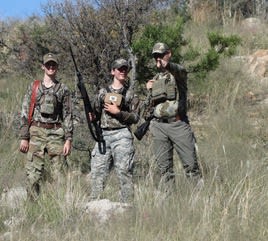 three young hunters in camo standing on a brushy hillside with a rifle