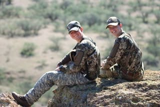 two boys in camo sitting on a rock, smiling back at the camera