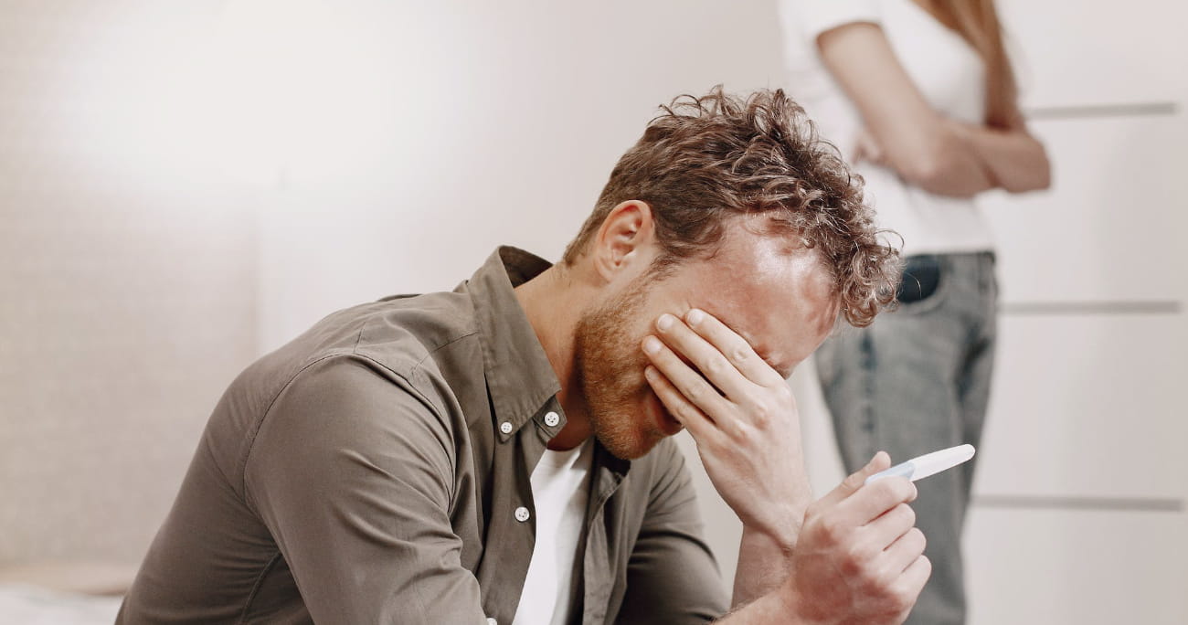 man sitting and crying holding a pregnancy test with his wife in the background