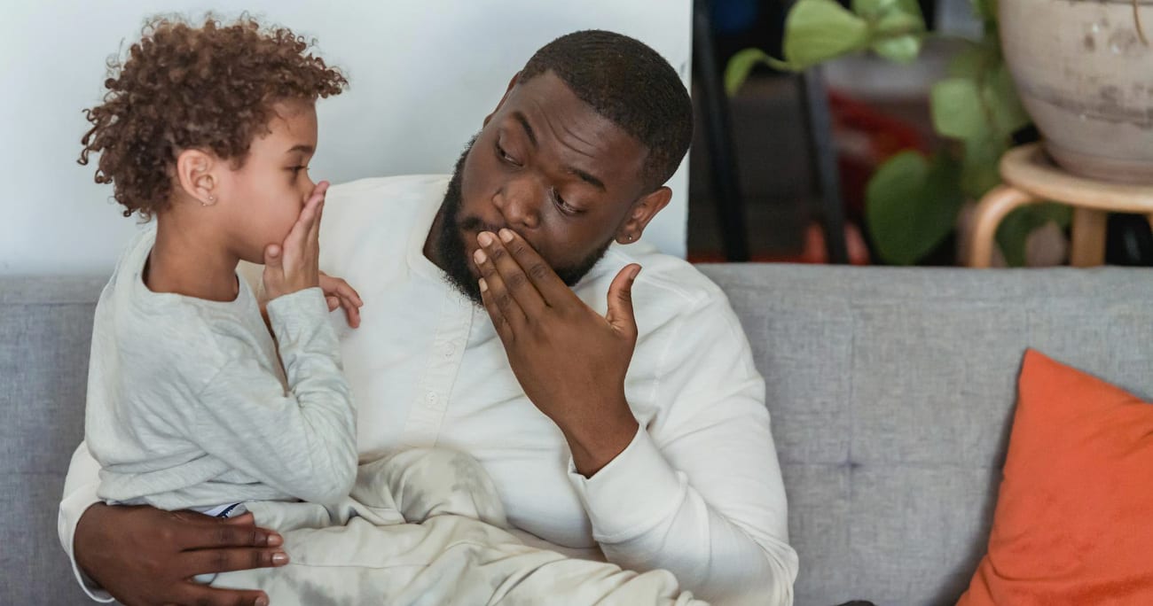 Black father with son covering mouth with hand on sofa