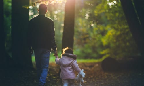A dad walking through the woods holding his young daughter’s hand