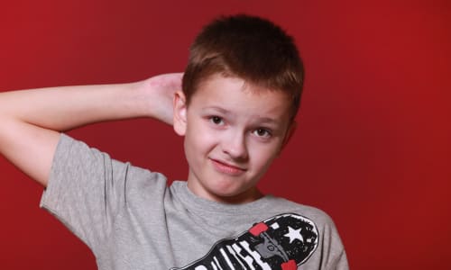 boy with a questioning face standing and holding one hand behind his head and looking at the camera