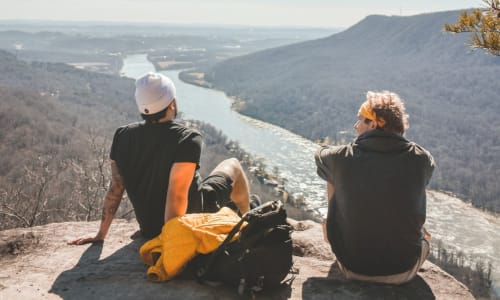 two guy sitting and talking overlooking a river in a valley below