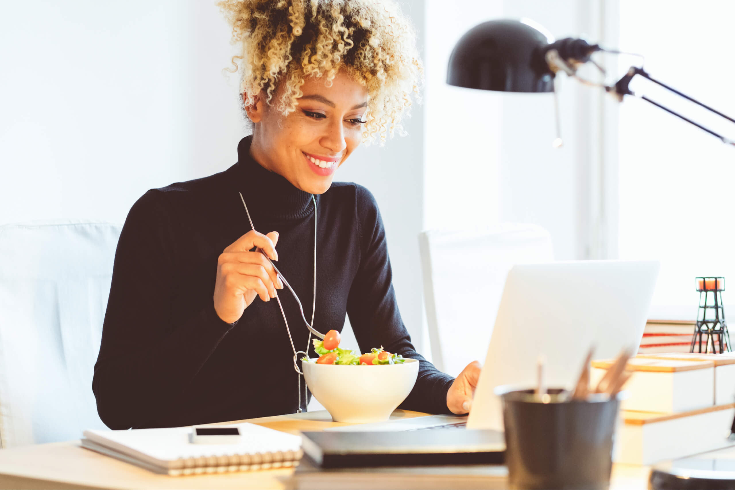 Woman smiling eating a salad while looking at her laptop screen.