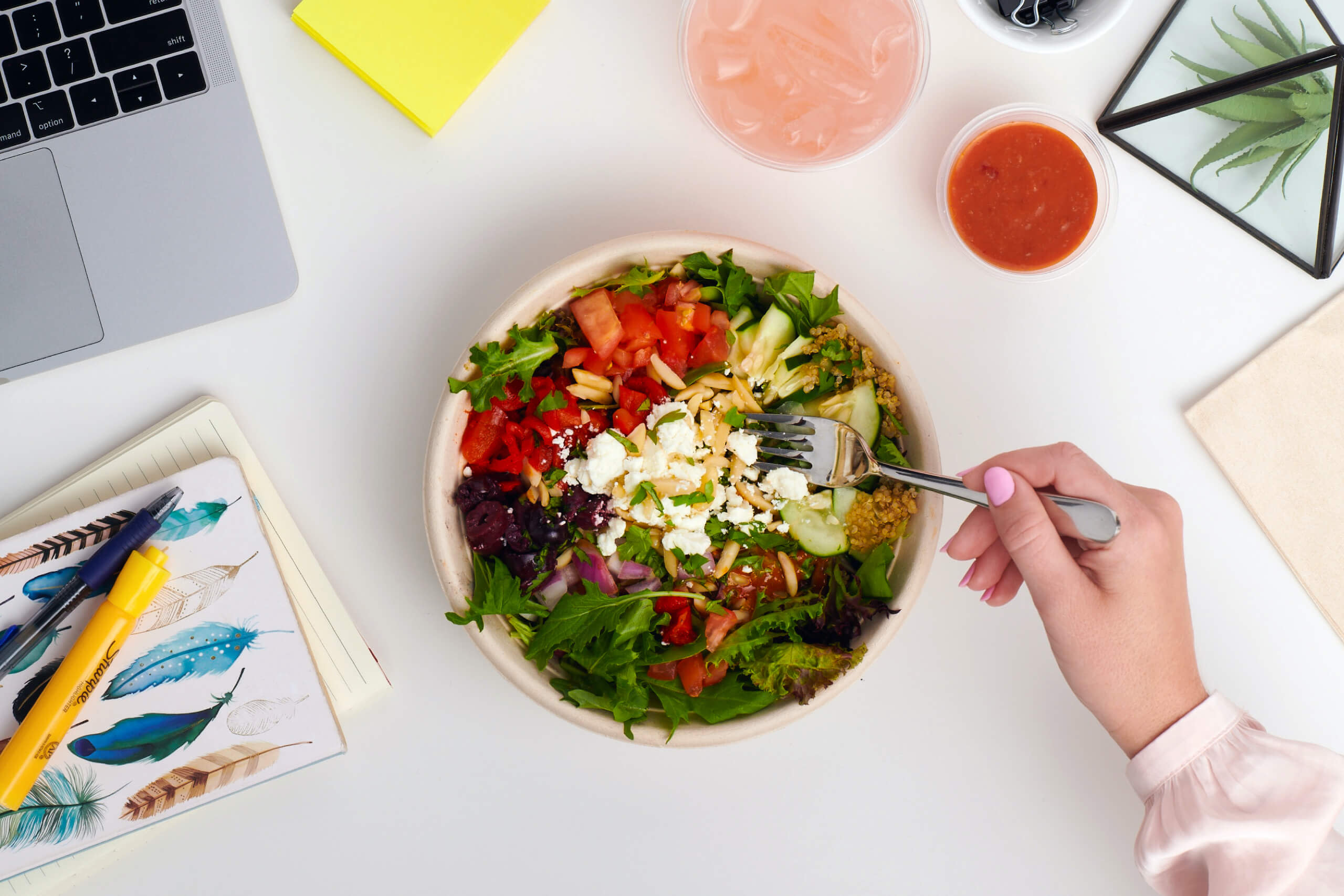 A salad on a desk with a female hand putting a fork in it to eat.