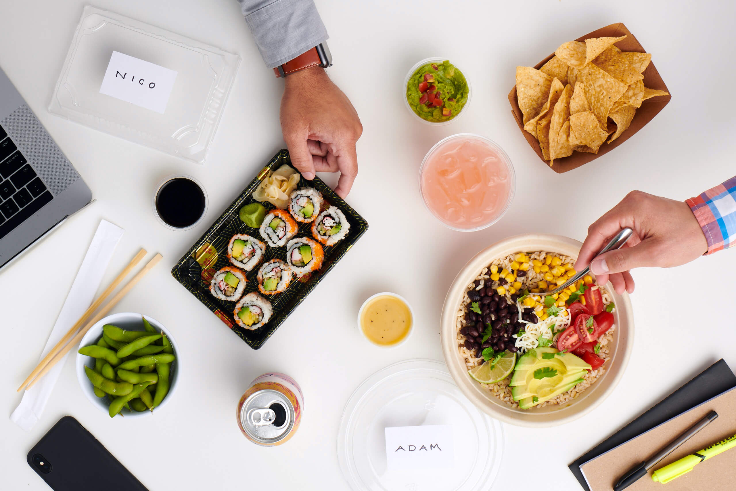 White desk with sushi, chips and guacamole, a soda can, a glass of pink lemonade and a burrito bowl with multiple hands reaching for the food items.