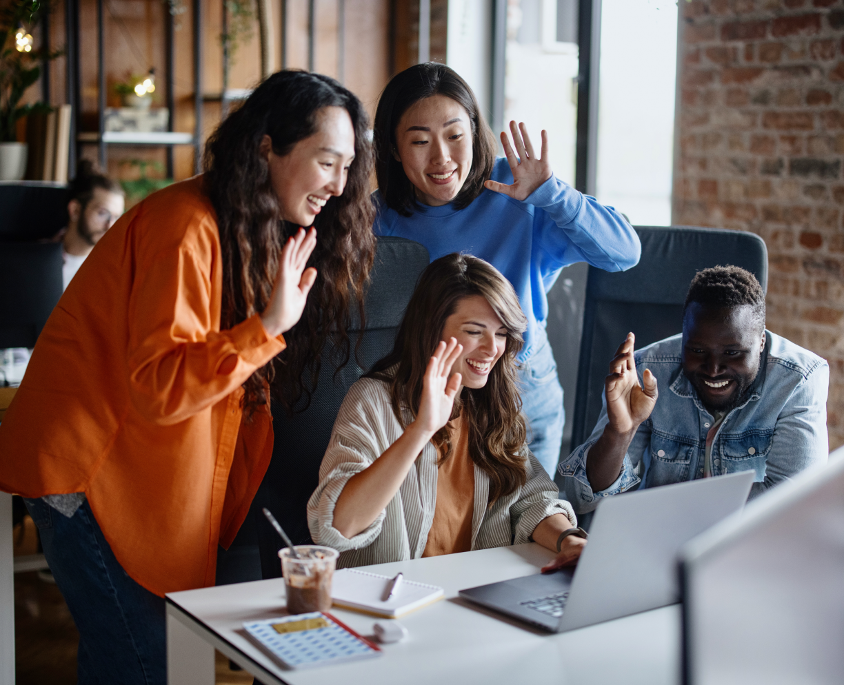 Three females and one male employee waving in front of a laptop on a video conferencing call.