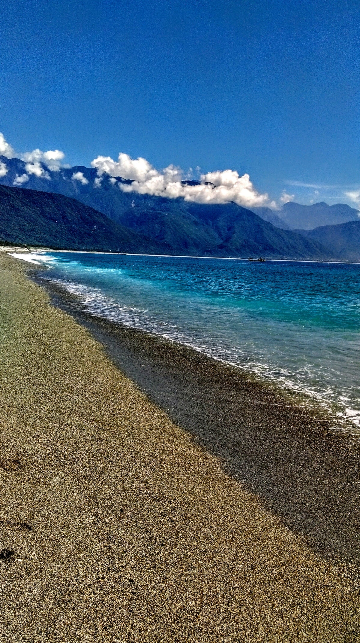 A curving grey pebble beach with turquoise water and green mountains beyond