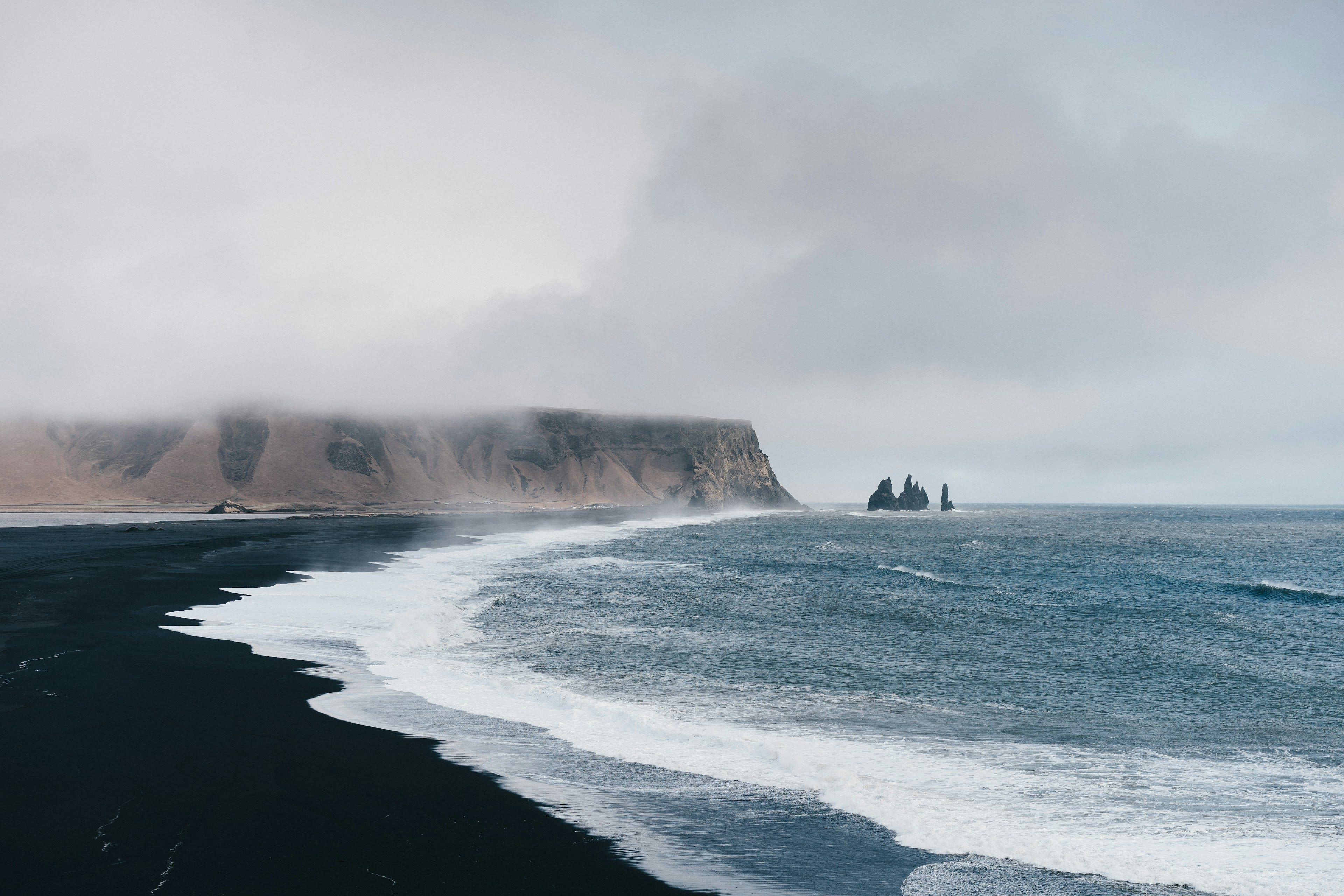 A curving black sand beach with white surf and basalt sea stacks in mist