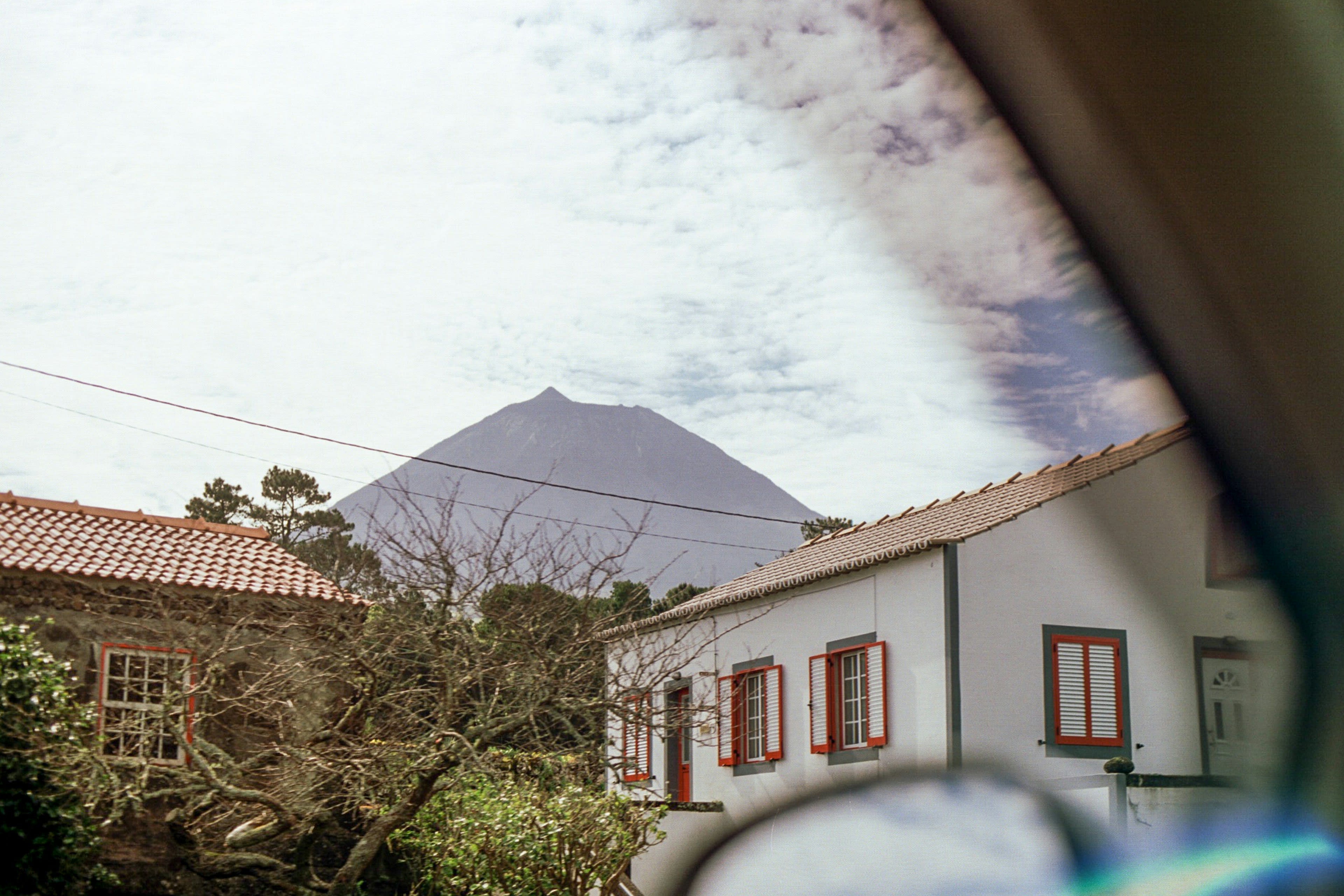 A white house with red shutters and a hazy volcano beyond, seen through a car window