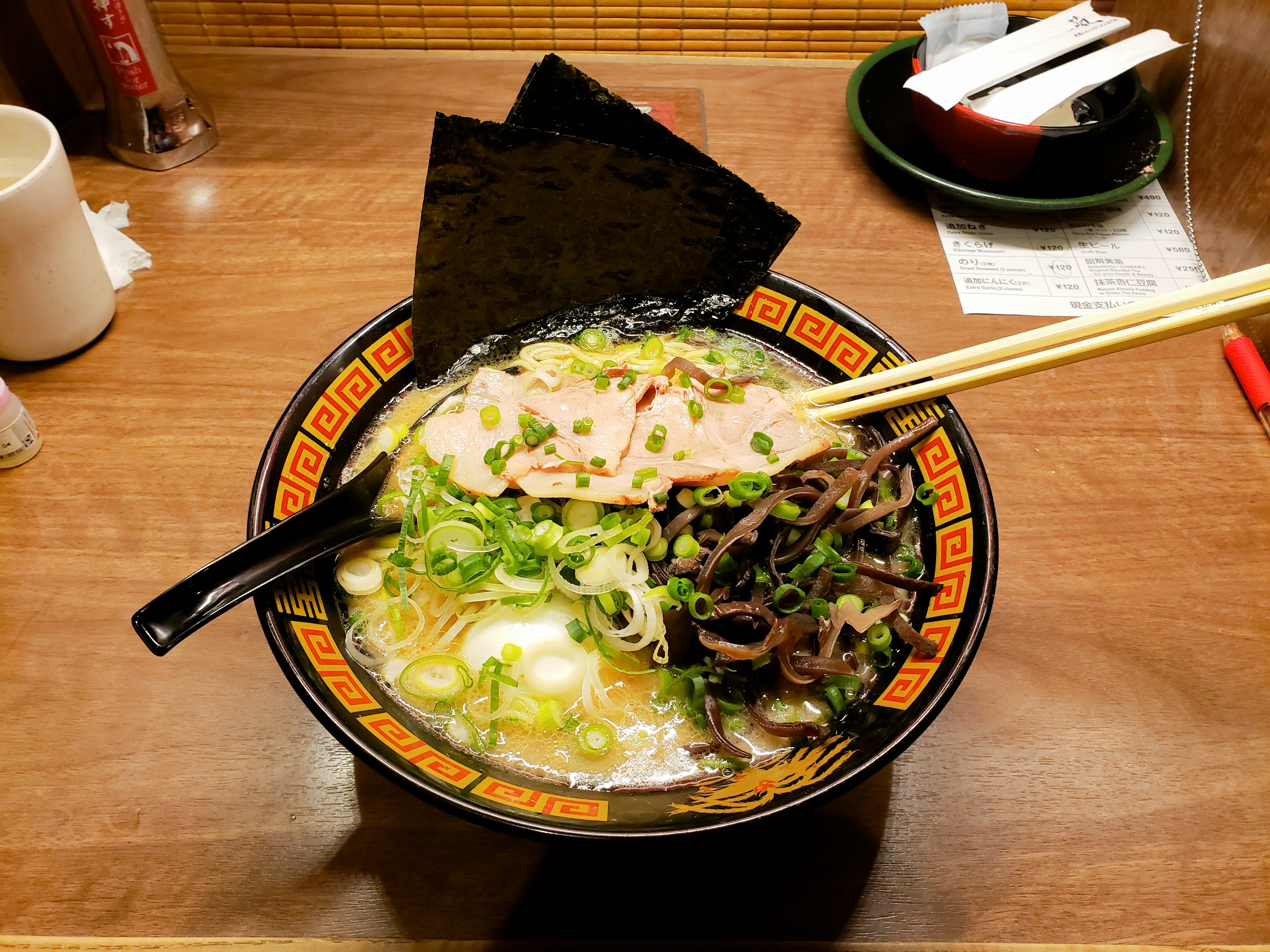 A bowl of ramen with sliced pork, spring onion and a sheet of nori on a wooden counter