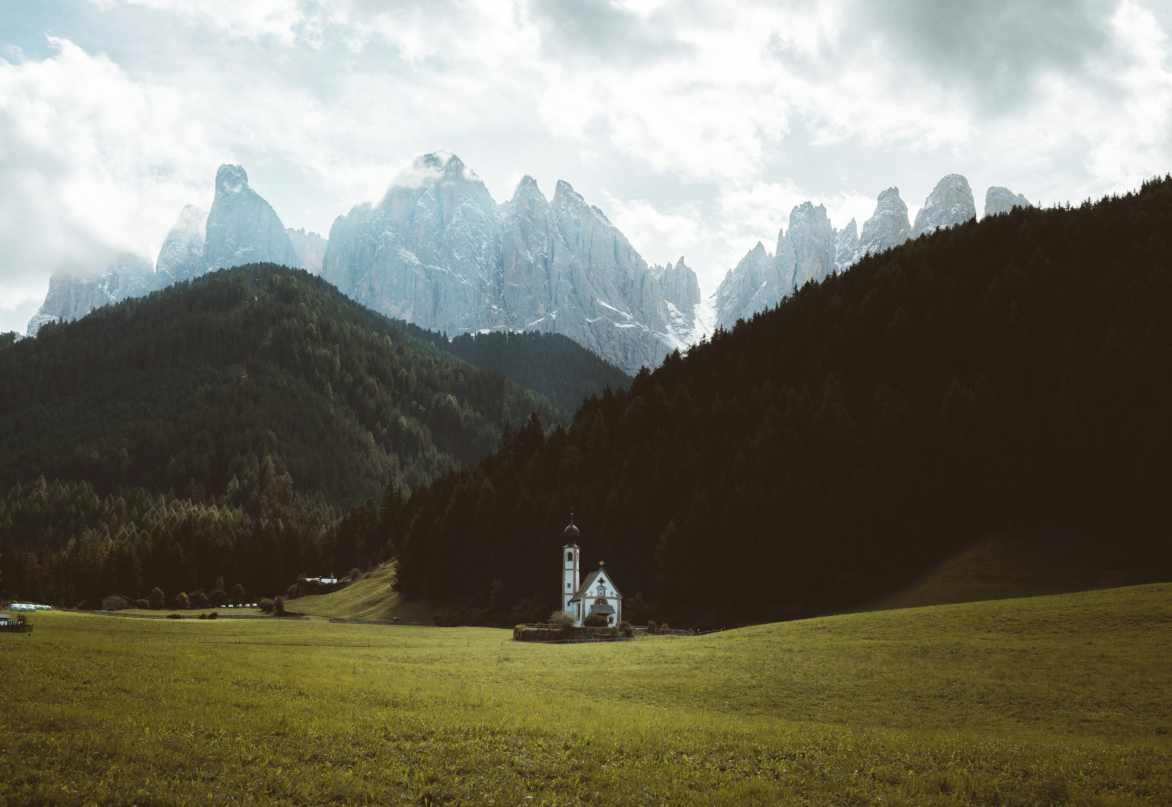 A small white chapel in a green meadow beneath the jagged Odle peaks and dark forest