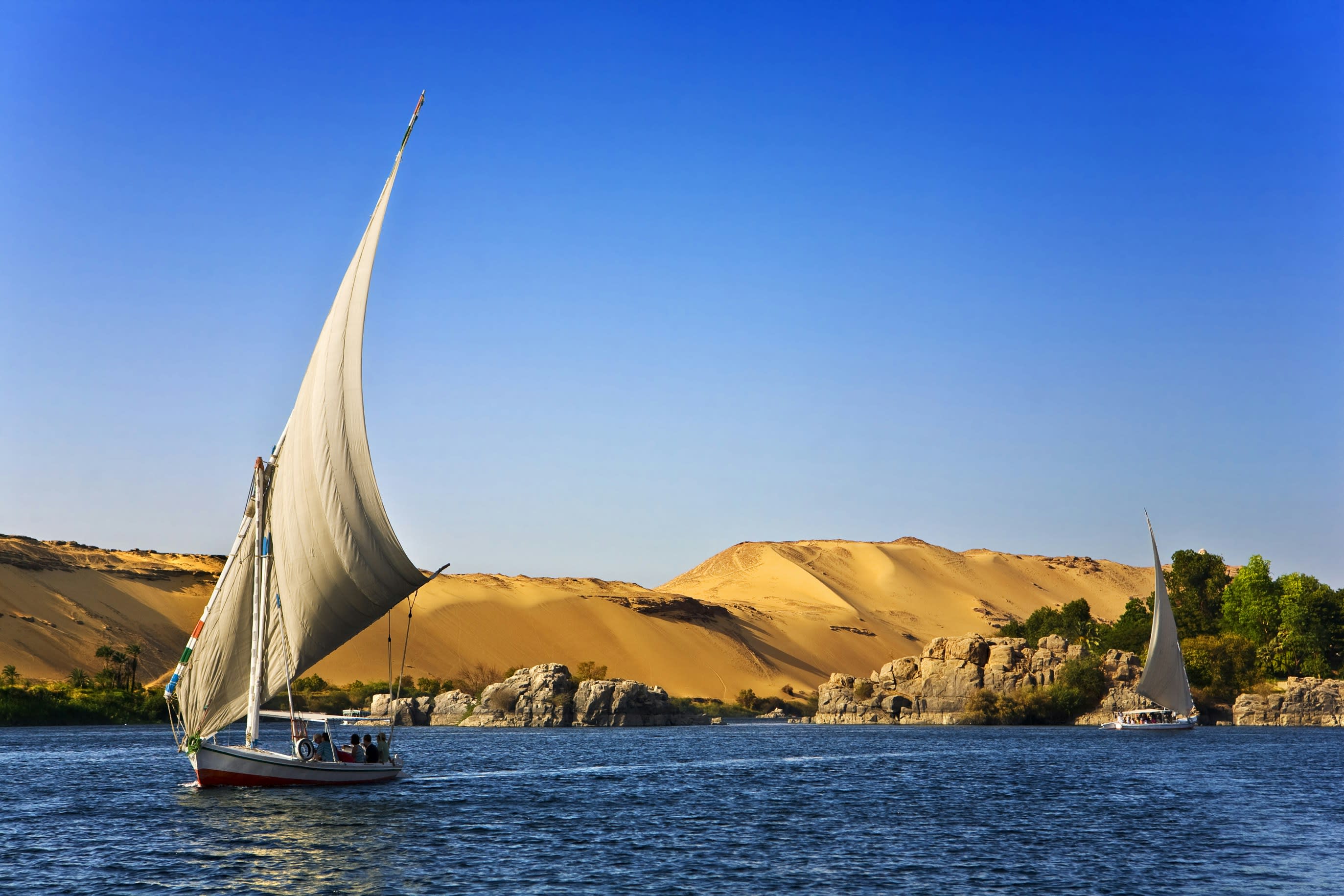 A white felucca sail against the dunes and blue water of the Nile at Aswan