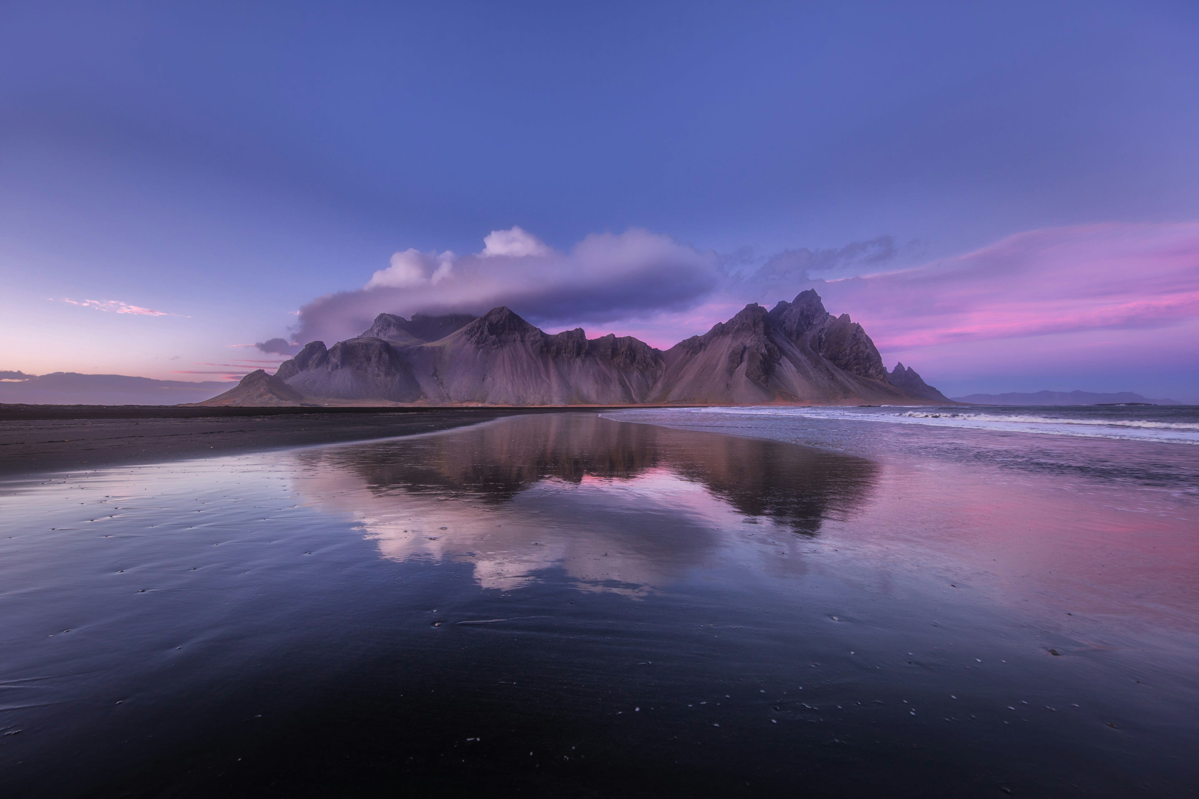 A jagged dark mountain reflected in wet black sand under a violet and pink sky