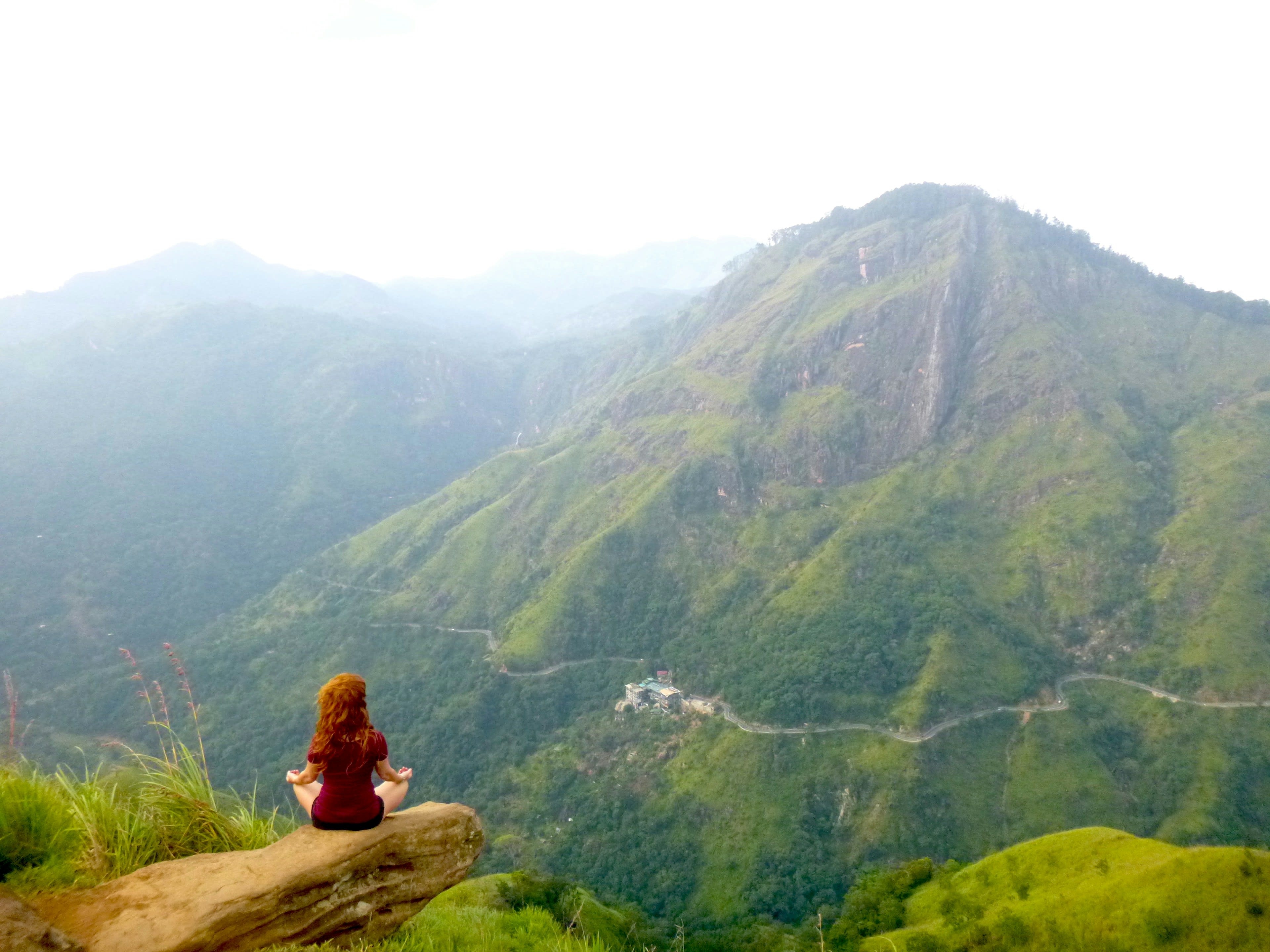 A person sitting on a grassy summit looking across a valley to a rocky peak