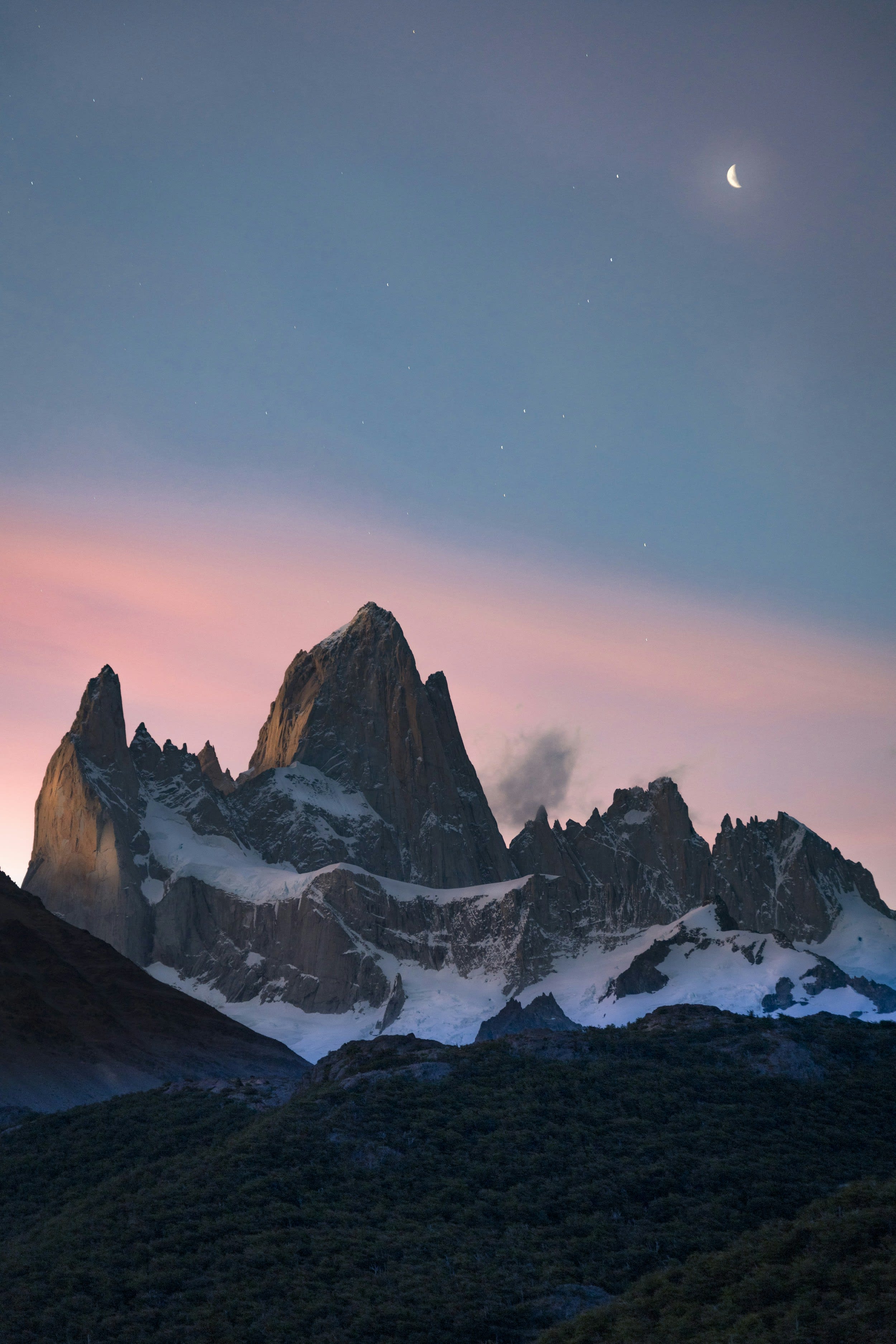 A dark granite peak against a pink dusk sky with a small moon above it